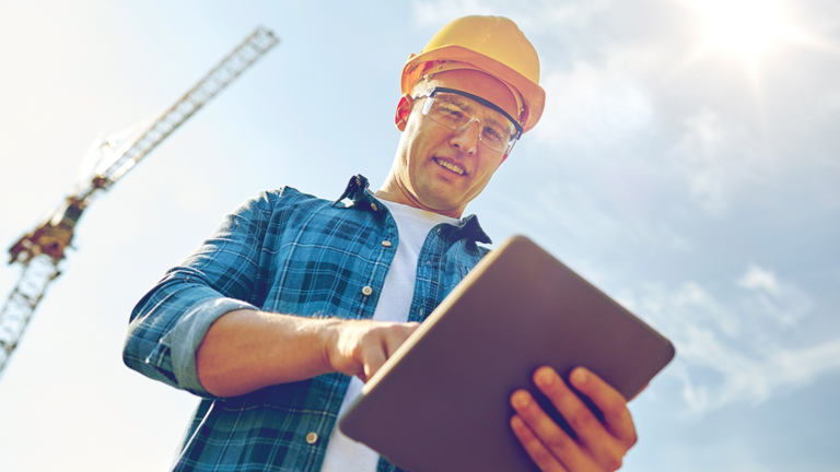 A man in a flannel shirt and hard hat working on a tablet on a construction site